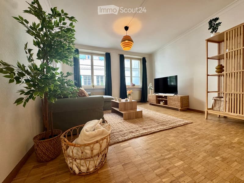 Modern living room with wooden flooring, large windows, a grey sofa, coffee table, TV, floor lamp, and decorative shelves.
