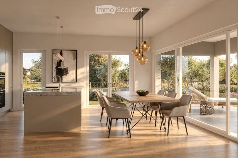 Modern kitchen and dining area with a central island, pendant lights, and a dining table with chairs. Large windows provide natural light and a view of the garden.