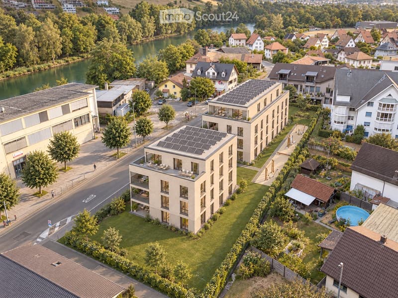 Aerial view of a modern residential complex with multiple buildings, solar panels on rooftops, a swimming pool, and surrounding greenery near a river.