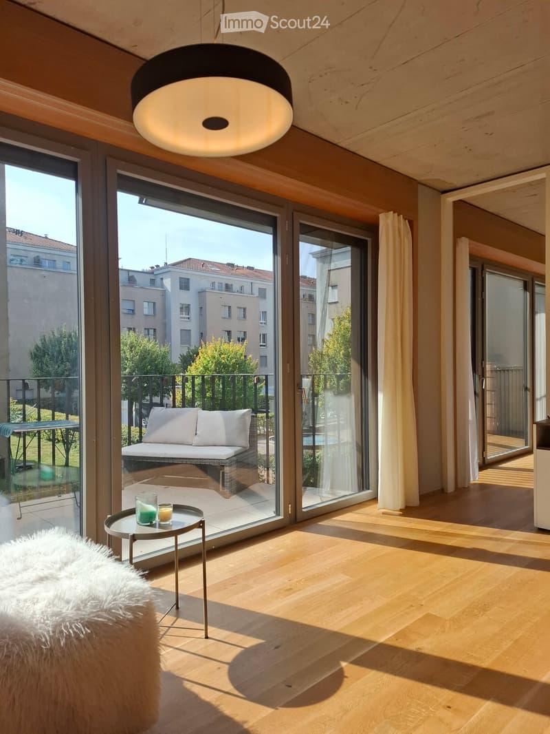 Bright living space with wooden flooring, large glass doors opening to a balcony, a small round table with two cups, and a fluffy white rug.