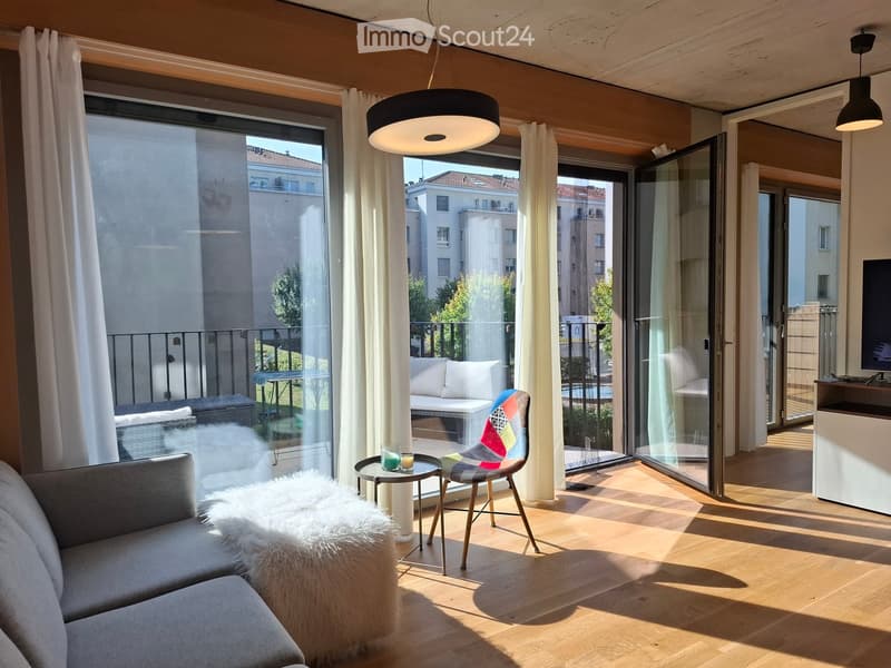 Modern living room with wooden flooring, large windows, and a balcony. Features a grey sofa, colorful chair, small round table, and decorative fur rug.