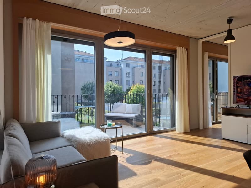 Modern living room with wooden flooring, large glass doors leading to a balcony, sofa, coffee table, ottoman, and a small table with drinks.