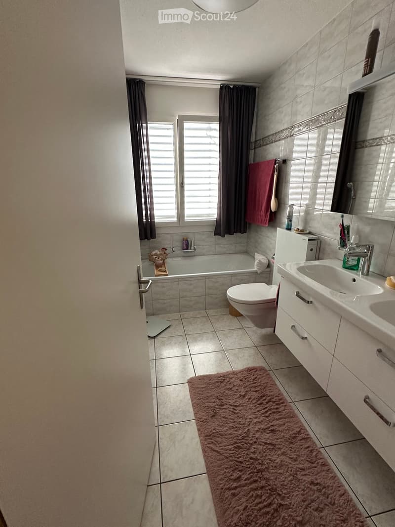 Modern bathroom with a bathtub, toilet, sink, mirror, and a pink towel on the wall. The room features tiled walls and floor, a window with dark curtains, and a fluffy pink rug on the floor.