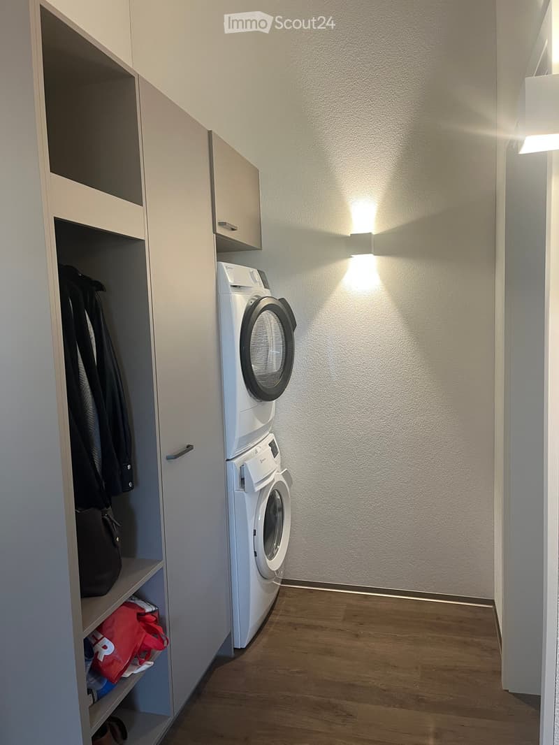 A small, modern laundry area with a white washing machine, a built-in wardrobe with hanging clothes, and a handbag on the shelf.