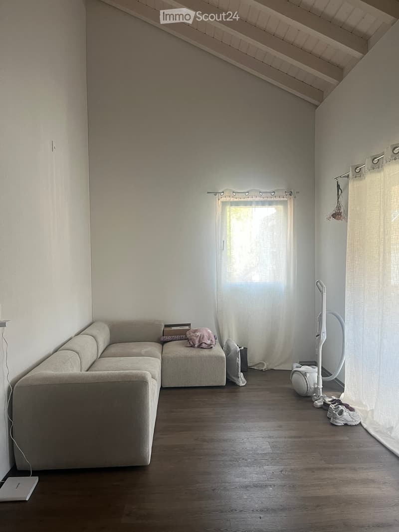 A modern living room with a sectional sofa, a laptop on the floor, a vacuum cleaner, and shoes near the window. The room features wooden flooring and large windows with curtains.