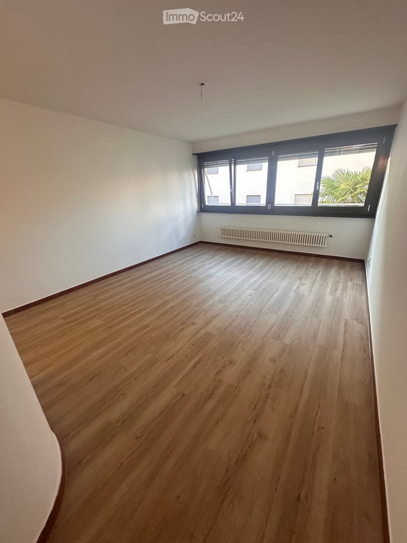 Empty room with wooden flooring, white walls, and a large window with blinds. A radiator is visible below the window.