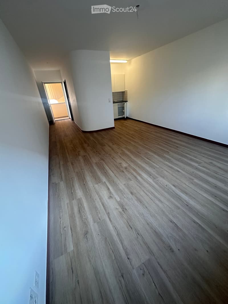 Empty hallway with wooden flooring and white walls, leading to an adjacent kitchen area.