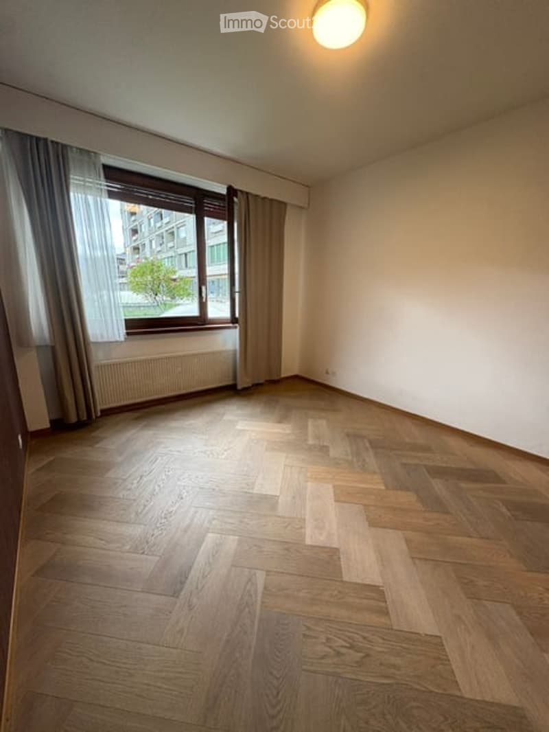 Empty room with wooden flooring, large window with curtains, radiator, and a ceiling light fixture.