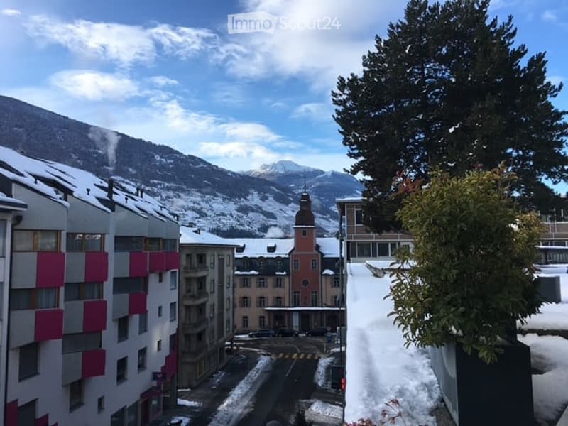 Balcony overlooking a snowy town with mountains in the background, featuring modern buildings and a church tower.