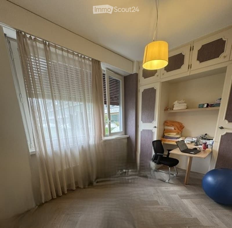 A well-lit room with wooden flooring, large windows covered by sheer curtains, a desk with a laptop, a chair, a cabinet, and a blue exercise ball on the floor.
