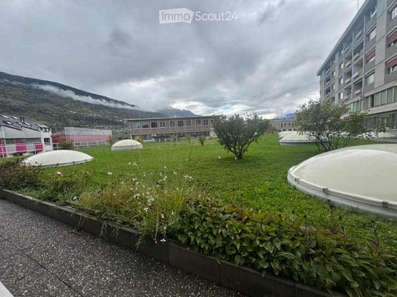 A well-maintained garden area with green grass, small trees, shrubs, and several dome-shaped structures in the background, with a modern building and mountains visible in the distance.