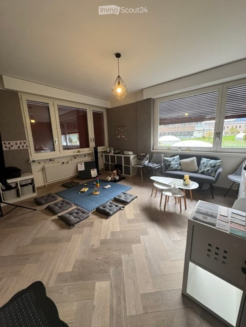 Bright living room with large windows, wooden flooring, and modern furniture including a sofa, coffee table, and several stools arranged on a blue mat.