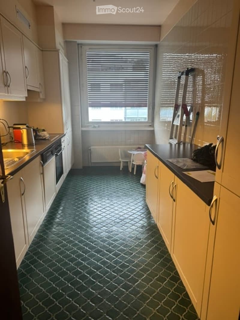 A compact kitchen with white cabinets, a dark green tiled floor, and a window with blinds. Visible appliances include a sink, fridge, and oven. A small stool and radiator are near the window.
