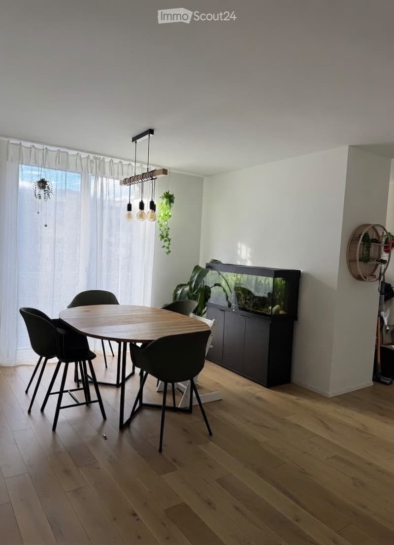 Modern dining area with a round wooden table, four black chairs, a large fish tank, pendant lights, and plants. The room features light wooden flooring and white walls.