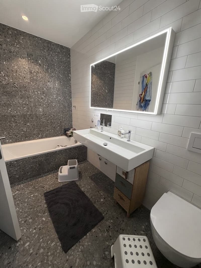 Modern bathroom with a double sink, large illuminated mirror, bathtub, toilet, and a small wooden cabinet. Includes a dark bathmat and a white stool.