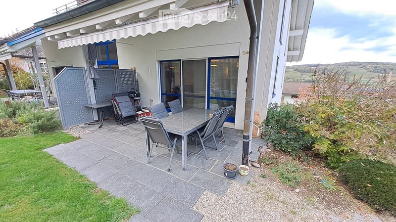 Outdoor patio area with a stone-tiled floor, a dining table with chairs, an umbrella, and surrounding greenery.