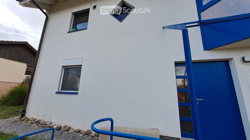 Modern two-story building with white exterior and blue window frames, featuring a raised ground floor entrance with blue railings.