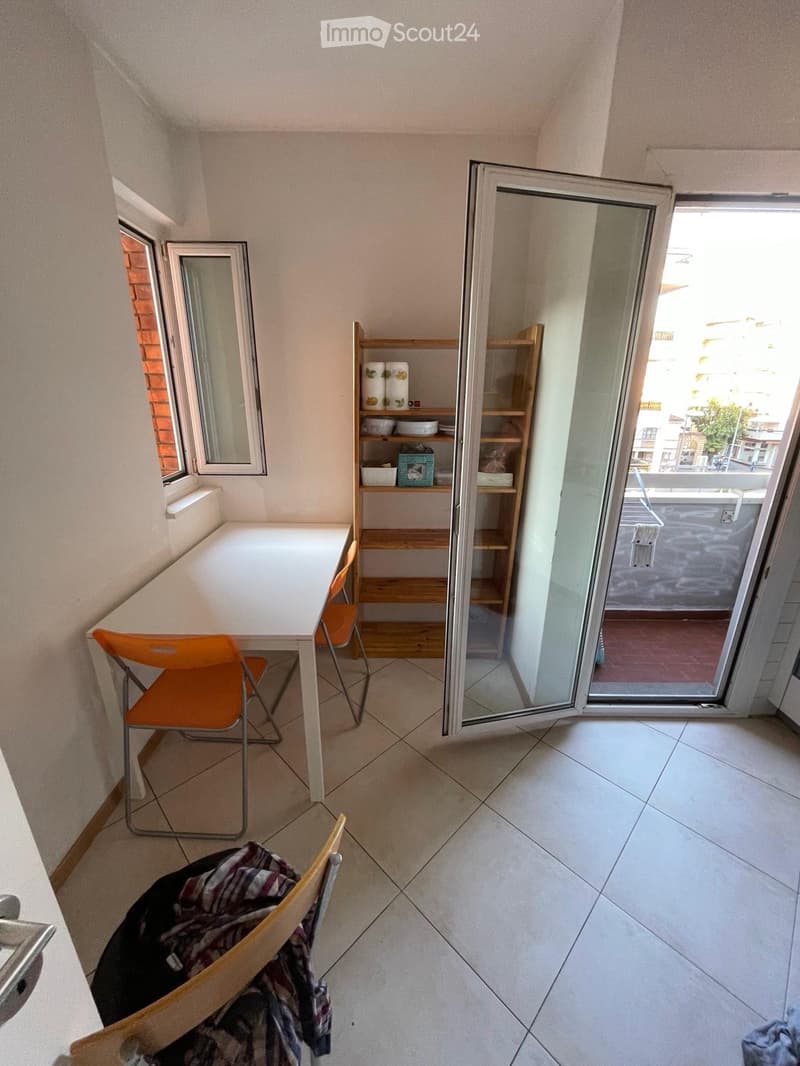 A small, furnished room with tiled flooring, a white table, two orange chairs, a wooden shelf with various items, and a sliding glass door leading to a balcony.