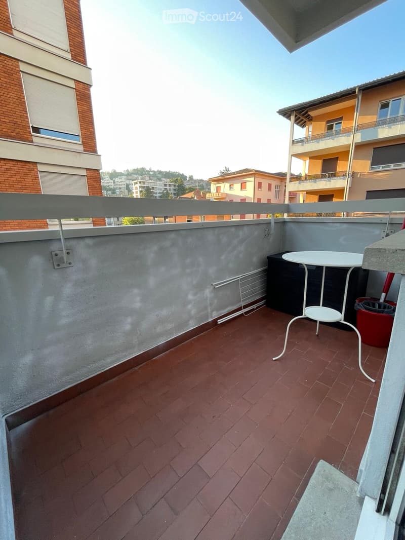 Balcony with red brick floor, white metal table, railing, and view of surrounding buildings.