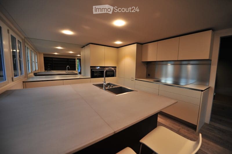 Modern kitchen with white cabinets, a central island, and a sink with a faucet. The space features built-in appliances, including an oven, and is illuminated by recessed lighting. There are two white stools near the island.