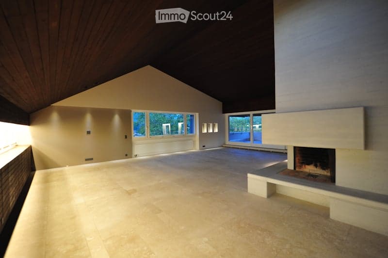 An empty room with a high, slanted wooden ceiling, large windows, and a fireplace with a built-in bench. The floor is tiled, and the walls are painted in neutral tones.