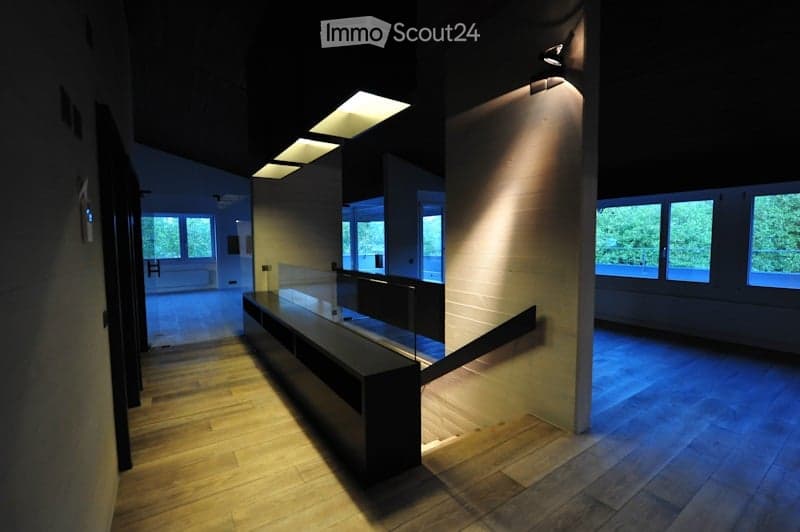 Modern hallway with wooden flooring, recessed lighting, and large windows allowing natural light. Features a sleek black bench and a staircase leading upwards.