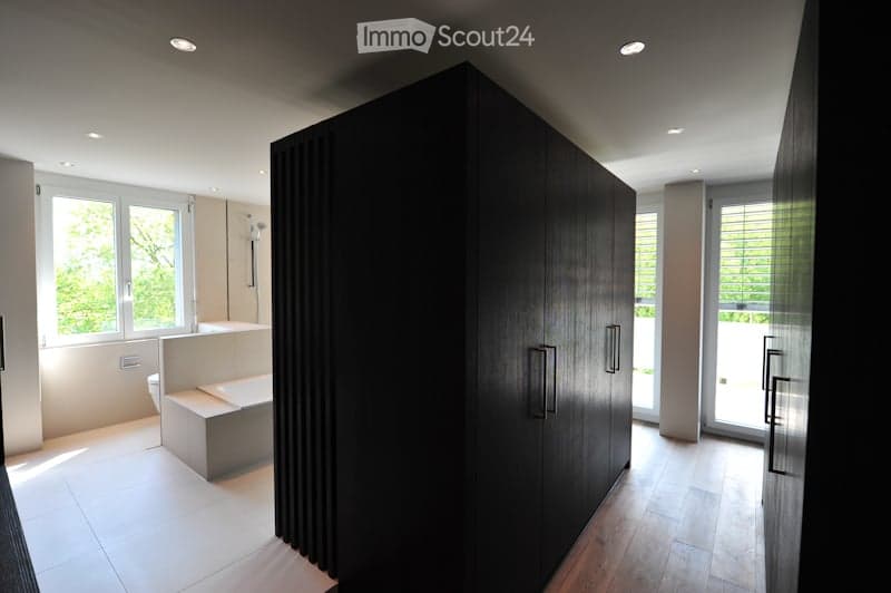 Modern bathroom with a large dark wooden cabinet, a bathtub, a toilet, and a sink. The space features large windows with blinds and recessed lighting.