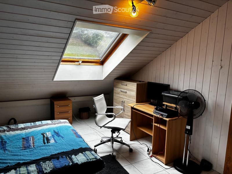 Cozy attic room with slanted wooden ceiling, skylight window, desk with computer and fan, office chair, bed with quilt, and small cabinet.