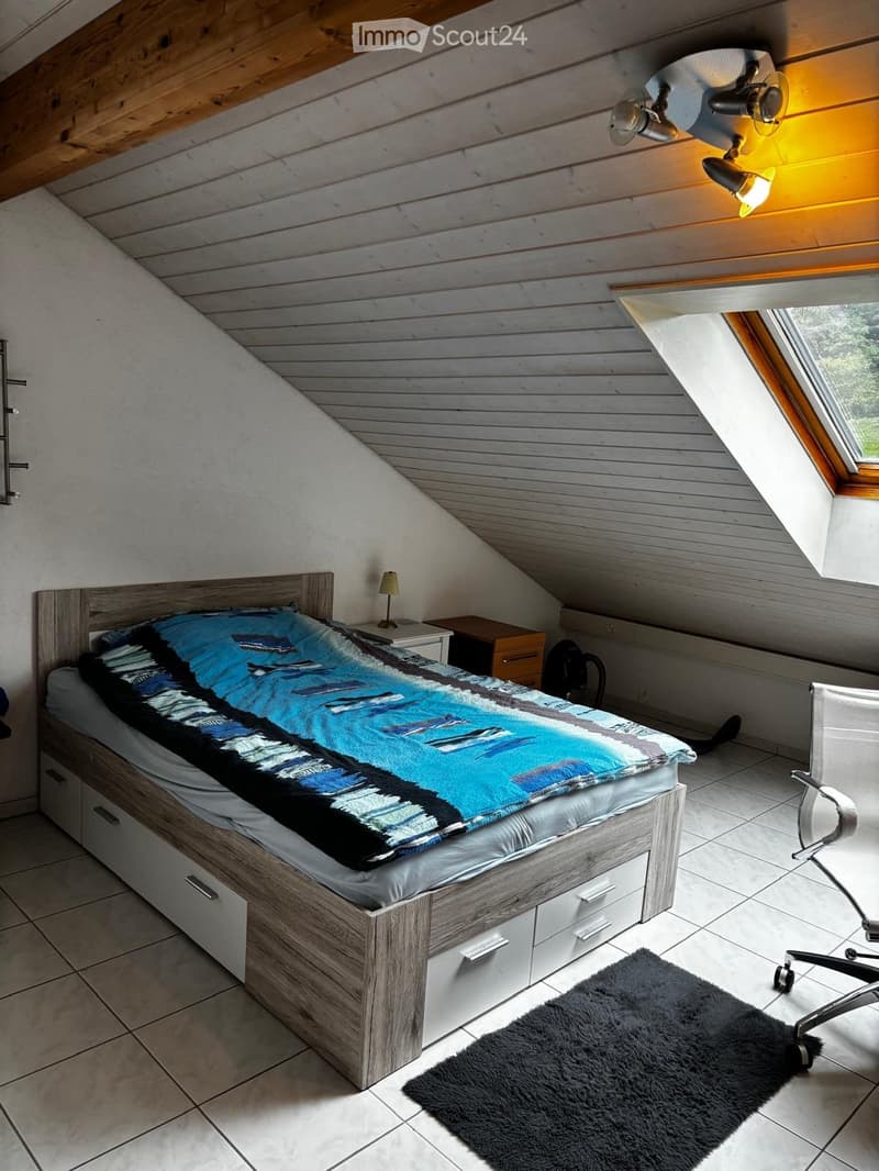 Cozy attic bedroom with wooden slanted ceilings, a single bed with a colorful blanket, a nightstand with a lamp, a chair, and a rug on the floor.