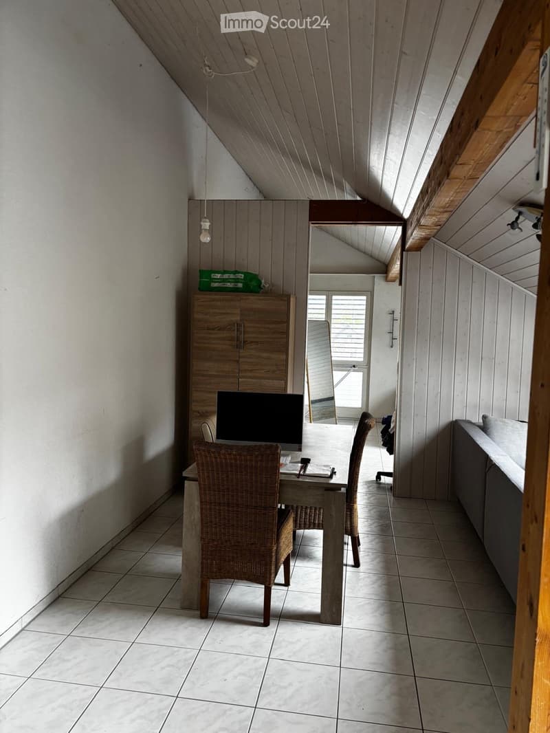 A cozy attic room with wooden walls and slanted ceilings, featuring a table with two wicker chairs, a sofa, a cabinet, and a screen on top of the cabinet.