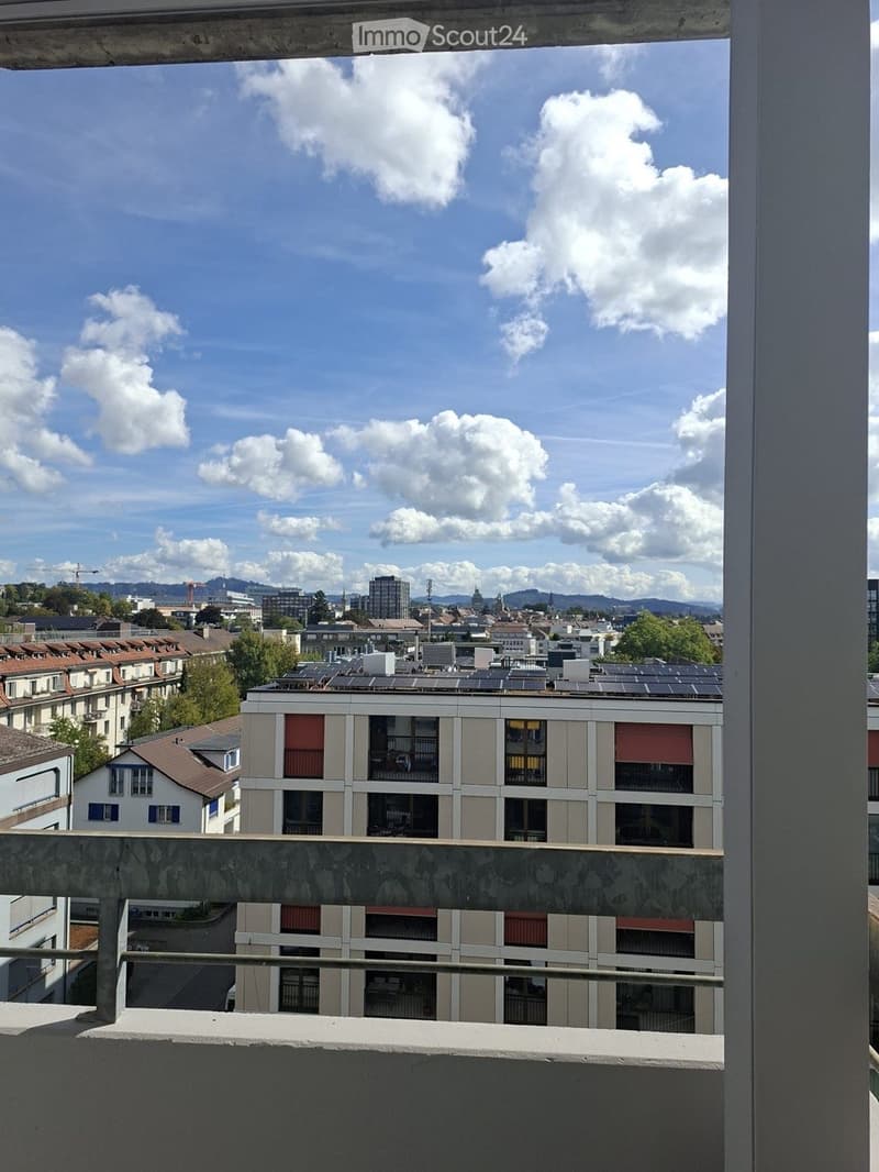 Balcony overlooking a cityscape with buildings, solar panels on rooftops, and a partly cloudy sky.