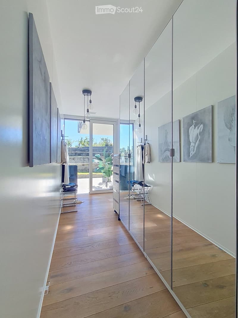 Modern hallway with wooden flooring, glass walls, and pendant lights. Features artwork on the walls and a view of an outdoor area through large glass doors.