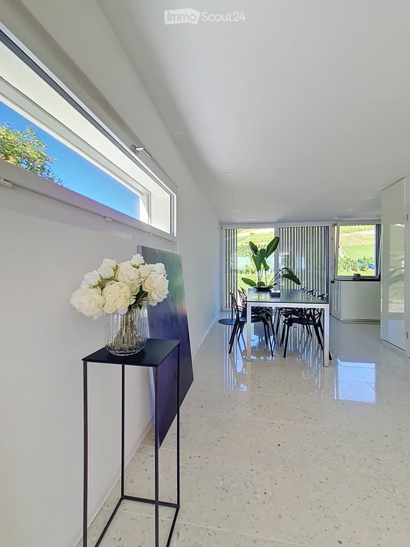 Bright, modern room with white walls, polished concrete floor, large window, and skylight. Features a small black stool with a vase of white flowers, a dining table with chairs, and a plant near the window.