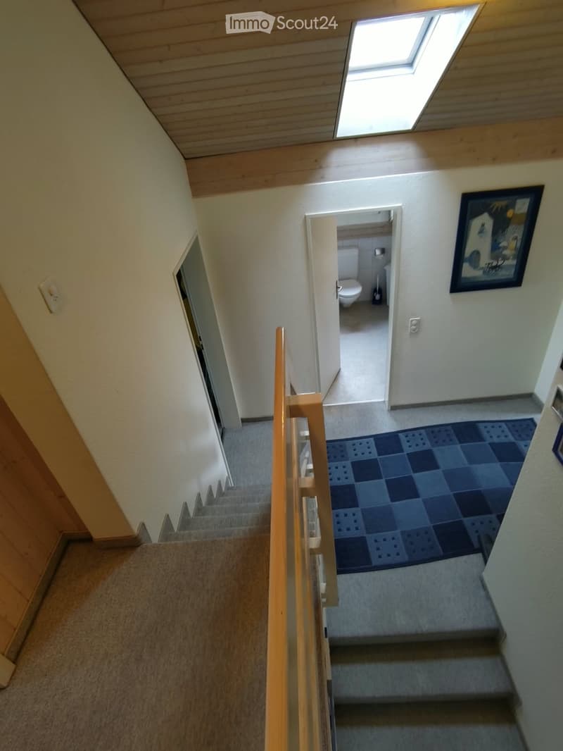 Staircase with wooden railing, carpeted steps, and a blue checkered rug at the landing. An open door leads to a bathroom with a toilet and a broom.