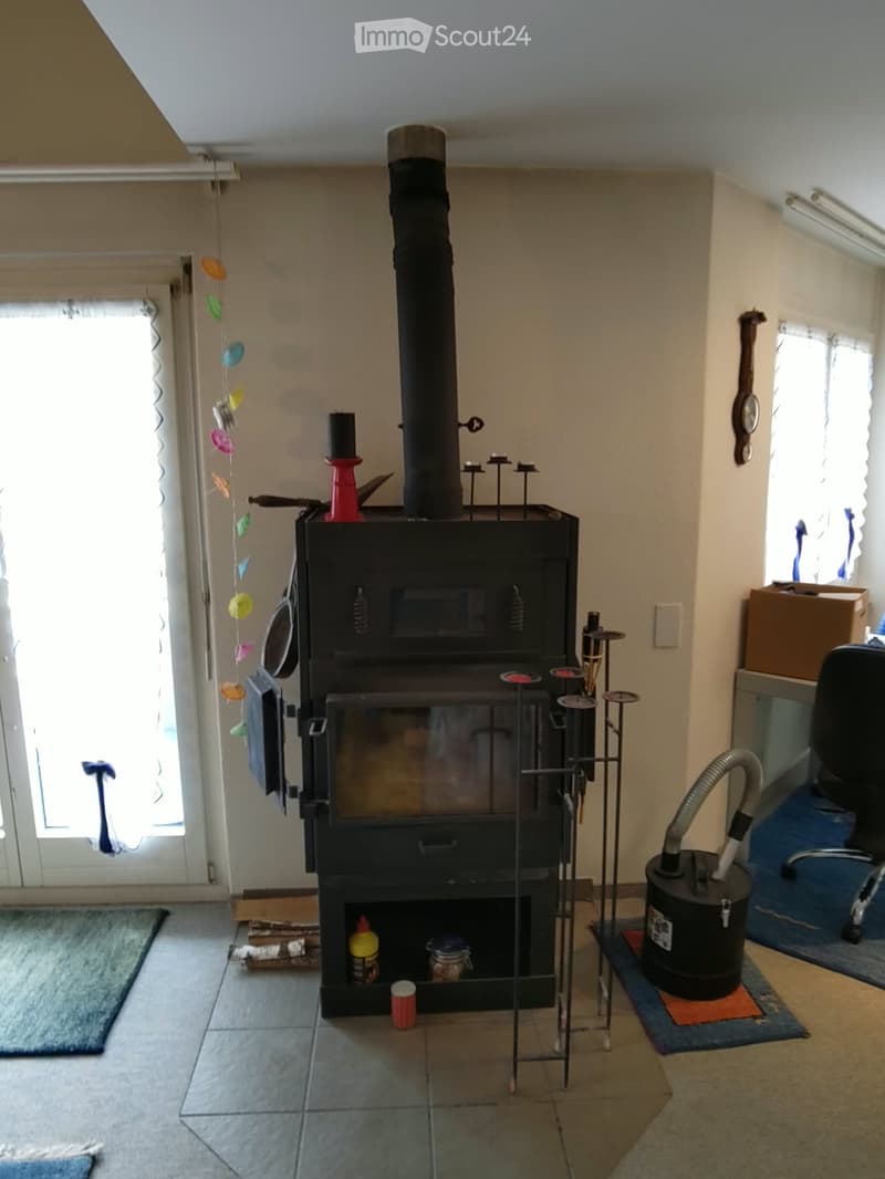 Living room with a central black wood-burning stove, surrounded by candles, tools, and a vacuum cleaner. The room has tiled flooring and large windows with blinds.