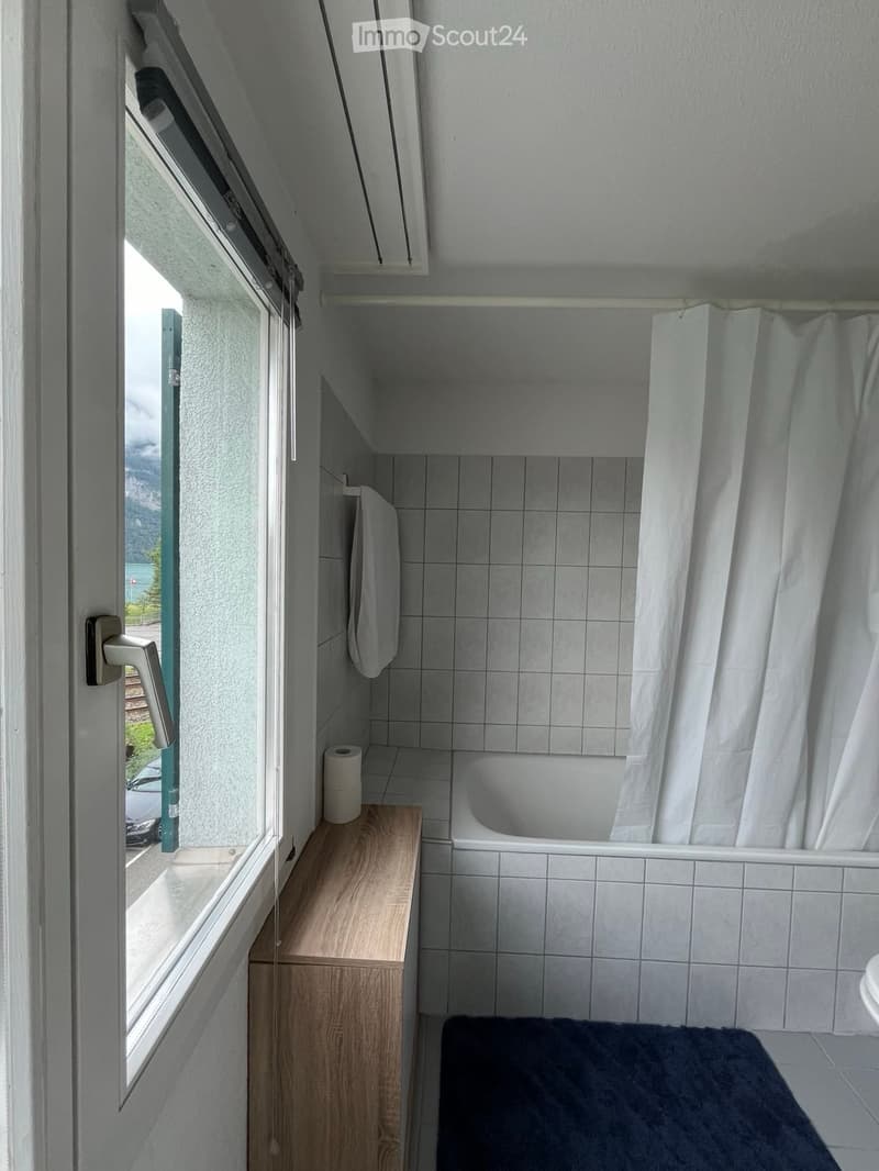 Compact bathroom with a bathtub, toilet, shower curtain, and a small wooden shelf holding a towel and toilet paper. A window offers a view of the outside.