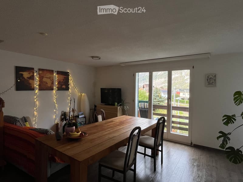 Bright living and dining area with wooden table, chairs, sofa, TV, and large windows leading to a balcony.