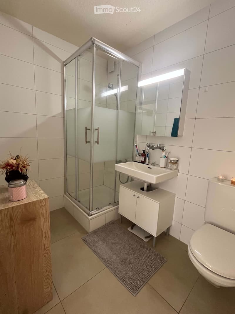 Modern bathroom with white tiles, a glass-enclosed shower, a white sink with a cabinet, a toilet, a mirror with built-in lighting, a small wooden shelf with a flower vase and candle, and a grey bath mat on the floor.