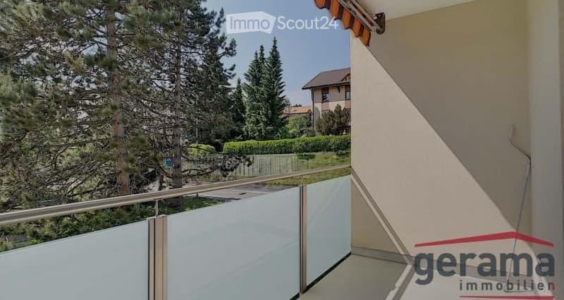Balcony with glass railing, overlooking a green area with trees and a neighboring house.