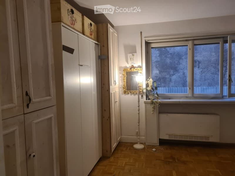 A small, well-lit room with wooden flooring, a large wardrobe, a mirror with a decorative frame, a floor lamp, a flower vase on a windowsill, and a radiator below the window.