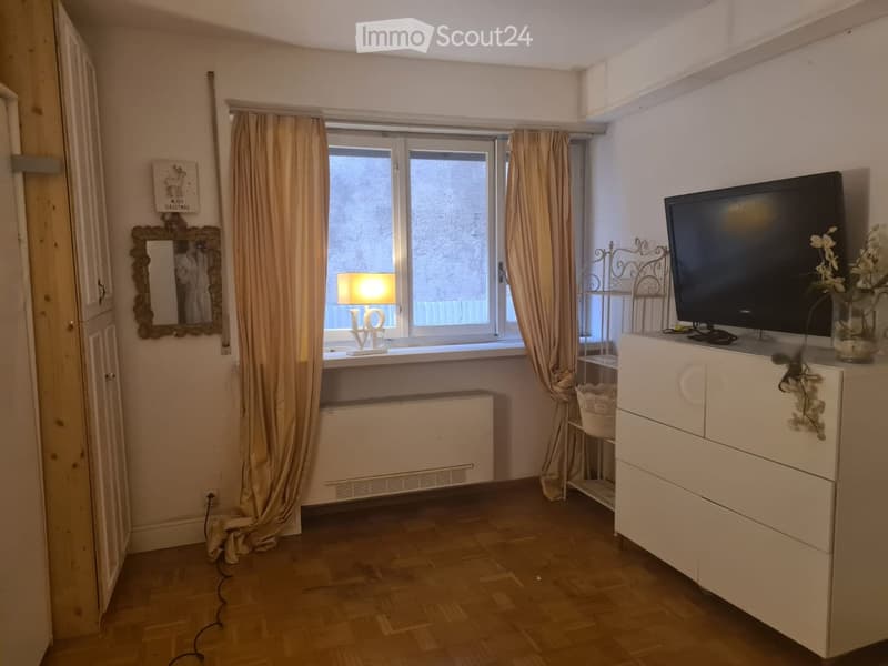 A furnished room with wooden flooring, a television on a white cabinet, a lamp on a windowsill, a decorative mirror, and a vase with flowers.