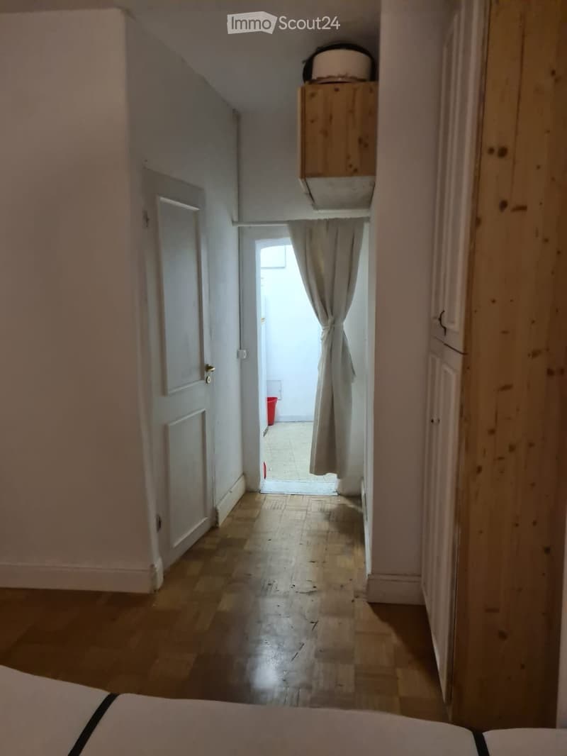 A narrow hallway with wooden flooring, white walls, and a small window at the end. There is a door on the left and a curtain on the right side.