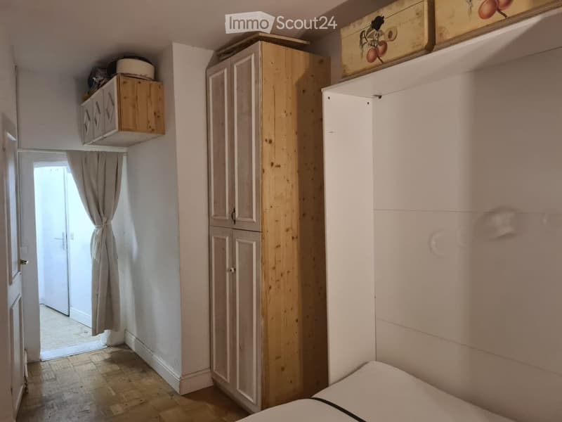 Bedroom with wooden floor, white walls, large wardrobe, small cabinet, and a curtained window leading to another room.