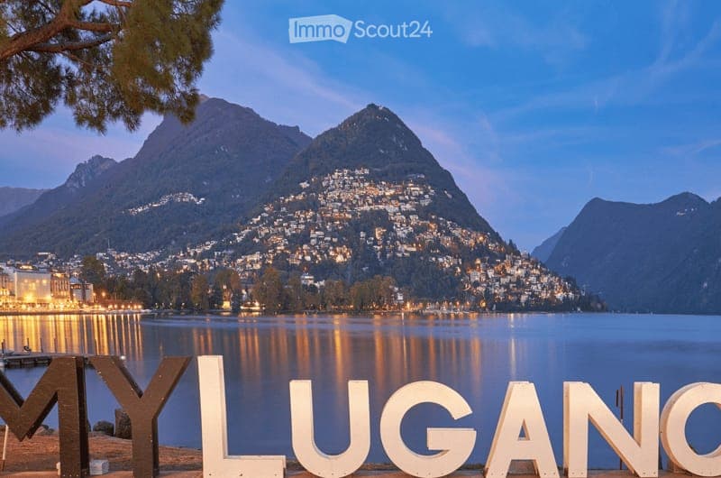 Scenic view of Lake Lugano with illuminated town and mountains in the background, featuring large white letters spelling "MY LUGANO" in the foreground.