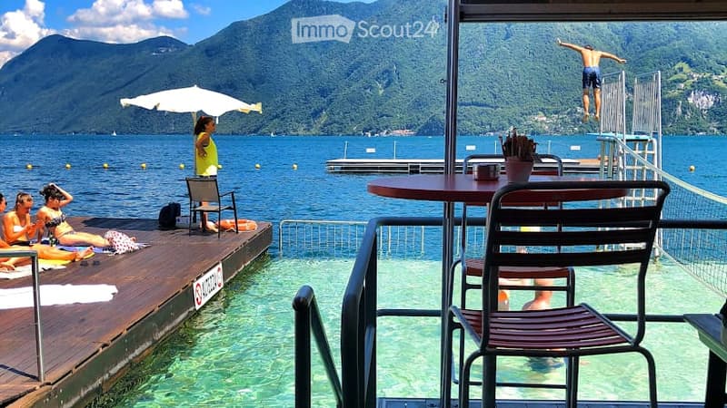 Outdoor area by a lake with wooden deck, lounge chairs, umbrella, and a diving board with a person jumping into the water. Scenic mountain view in the background.