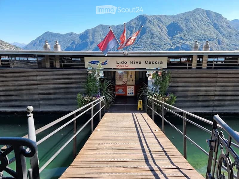 A wooden boardwalk leads to a building labeled "Lido Riva Caccia" with flags and a scenic mountain view in the background.