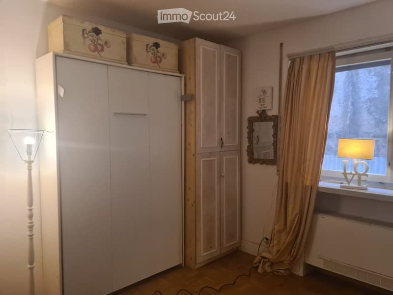 A furnished room with a large wardrobe, a mirror, a table lamp on the windowsill, a floor lamp, and curtains covering the window.