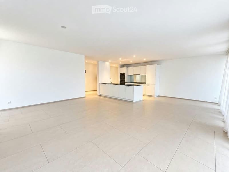 Empty room with white walls, tiled floor, and a white kitchen area with cabinets and appliances.