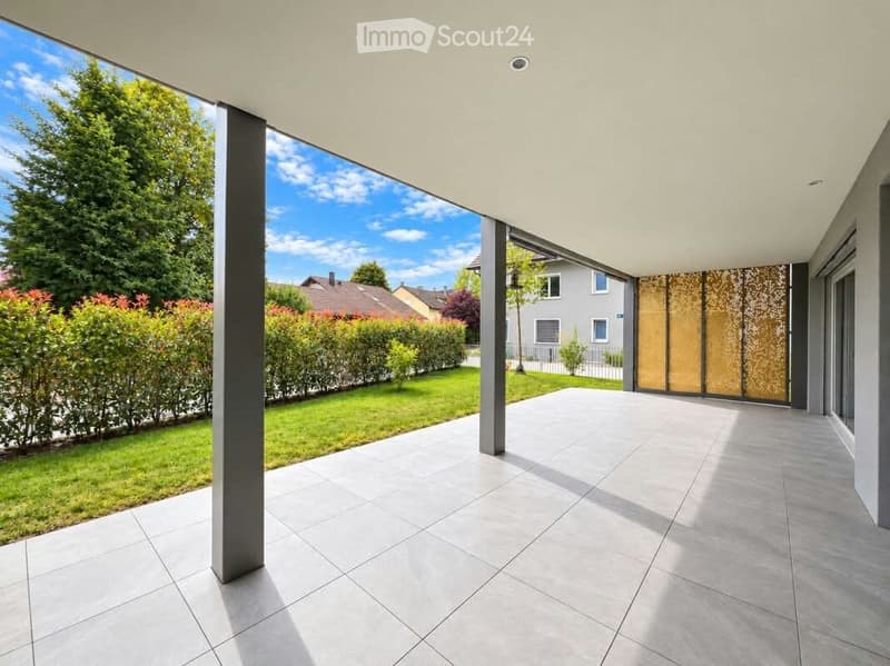 Modern outdoor patio with large tiled floor, grey columns, and a view of a well-maintained garden and neighboring houses.
