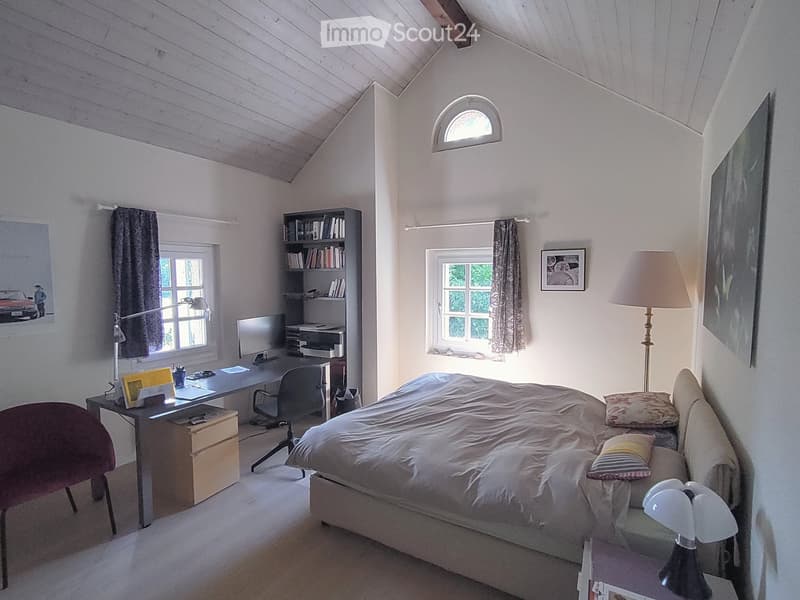 Cozy bedroom with a large bed, desk, bookshelf, and two windows with curtains. The room features a slanted wooden ceiling and warm lighting.